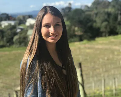 A woman with long brown hair stands outdoors in a rural setting, smiling at the camera. The background features a green field and blurred trees under a partly cloudy sky. She is wearing a blue top.