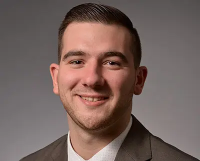 A person with short, dark hair smiles while wearing a grey suit and white shirt against a plain background.