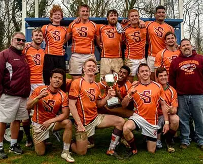 A rugby team poses outdoors with a trophy, wearing orange jerseys with a large "S" on the front. Two coaches in maroon stand on either side of the group. The team looks happy and triumphant, celebrating their victory.