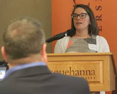 A woman in glasses speaks at a podium with a microphone. The podium has "Susquehanna University" written on it. An audience member is partially visible in the foreground. An orange banner with text is in the background.
