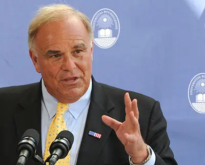Man in a suit and yellow tie speaks at a podium with microphones. He gestures with his right hand. The background features a blue banner with a white emblem depicting an open book and a sun.