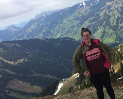 A person stands on a grassy hillside with a backdrop of lush, green mountains and a cloudy sky. They are wearing glasses, a brown jacket, and a maroon hoodie. The scene is serene and picturesque.