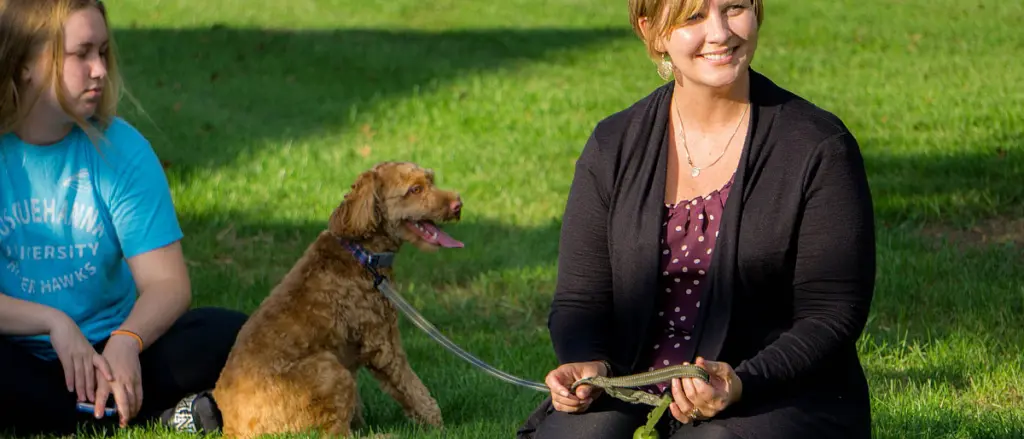 A woman sitting on the grass, smiling, while holding a book. Next to her, a girl in a blue T-shirt with "University" partially visible sits holding a brown dog on a leash. The dog is panting happily. The scene is sunny and relaxed.