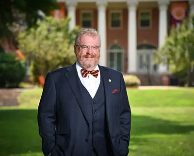 A man in a dark suit and bow tie stands on a grassy lawn with his hands in his pockets. There's a building with columns in the background, and trees are visible on either side. He is smiling at the camera.