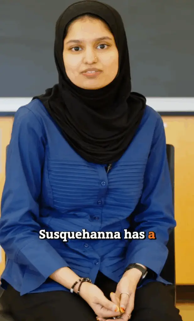 A person wearing a blue shirt and black headscarf is seated in a room with wooden panels and a chalkboard. They have a slight smile. The text on the image reads, "Susquehanna has a".
