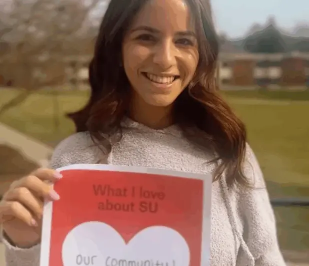 A person smiling, holding a red and white paper that says "What I love about SU" with "Our community!" written inside a heart. They are standing outdoors with a blurry campus background.