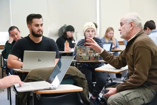 A classroom scene with a bearded older man talking to a younger man seated at a desk with a laptop open. Other students in the background are focused on their laptops and notebooks. The room has bright lighting and neutral-colored walls.
