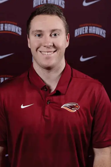 A person in a maroon polo shirt with embroidered logos stands smiling in front of a maroon backdrop with "River Hawks" and Nike logos.
