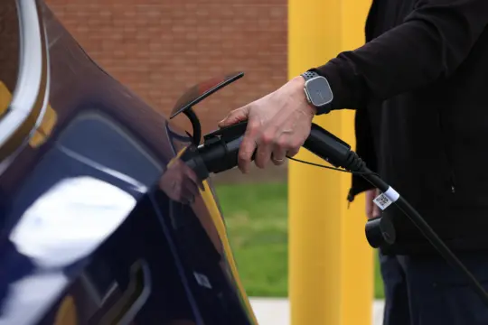 A person charges an electric car by plugging a charging cable into the vehicle. There is an emphasis on sustainability. The background shows a brick wall and a yellow post.