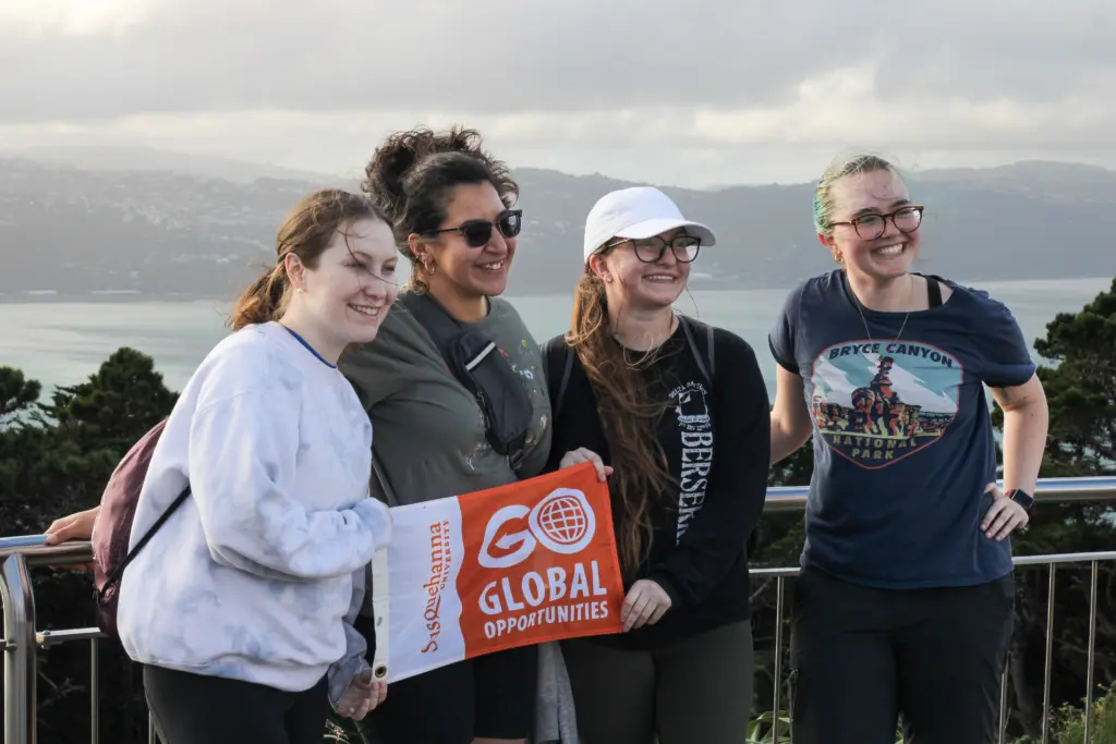 Four people stand together outdoors, smiling and holding a sign reading "Go Global Opportunities." They are on a lookout point with a scenic view of water and hills in the background.