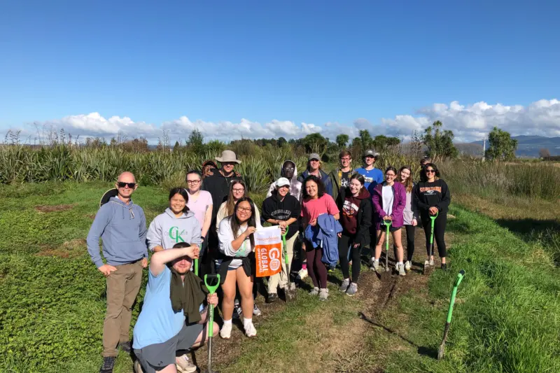 A group of people stand together outdoors in a grassy area under a blue sky. They are holding a "Go Slugs!" banner and appear ready for an activity, some holding shovels. Vegetation and fields are visible in the background.