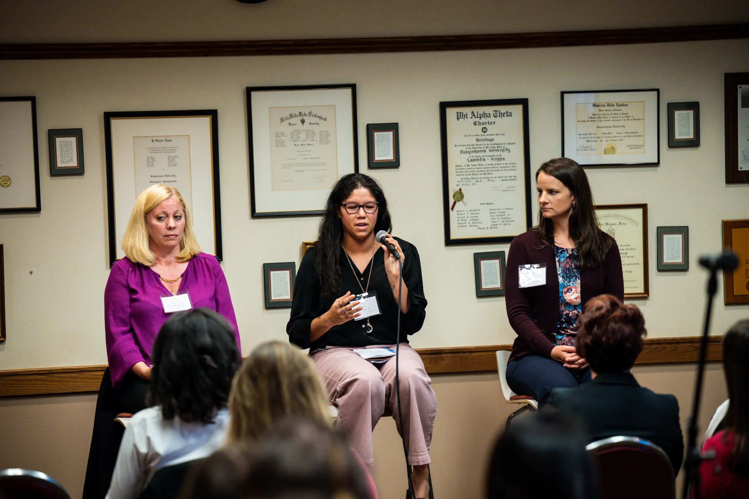Three women sit on stage in front of a wall with framed certificates, participating in a panel discussion. The woman in the center holds a microphone while speaking. An audience listens attentively.
