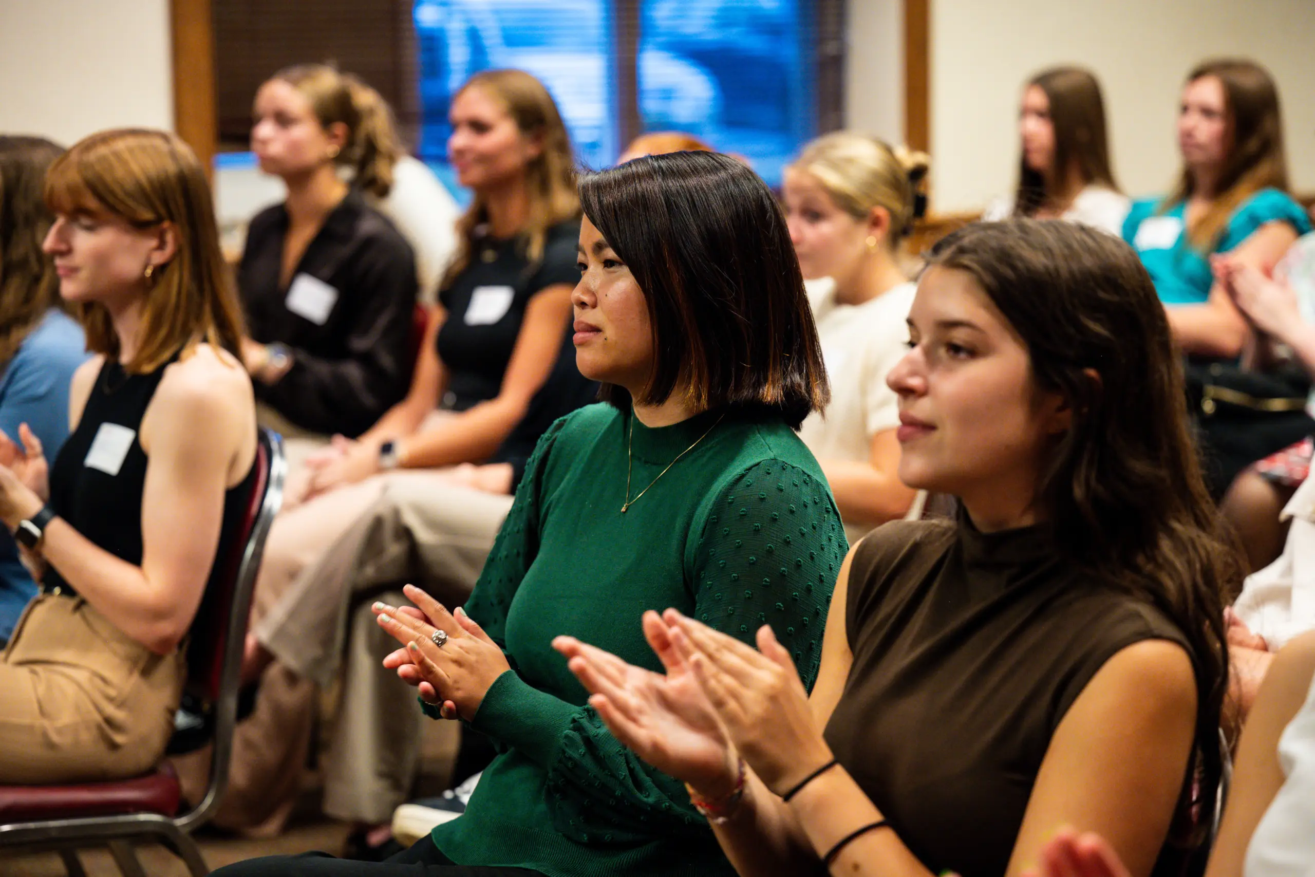 A group of people sitting in rows attentively clapping, with several women visible in focus. They appear to be attending an event or presentation in a room with natural light coming through a window.