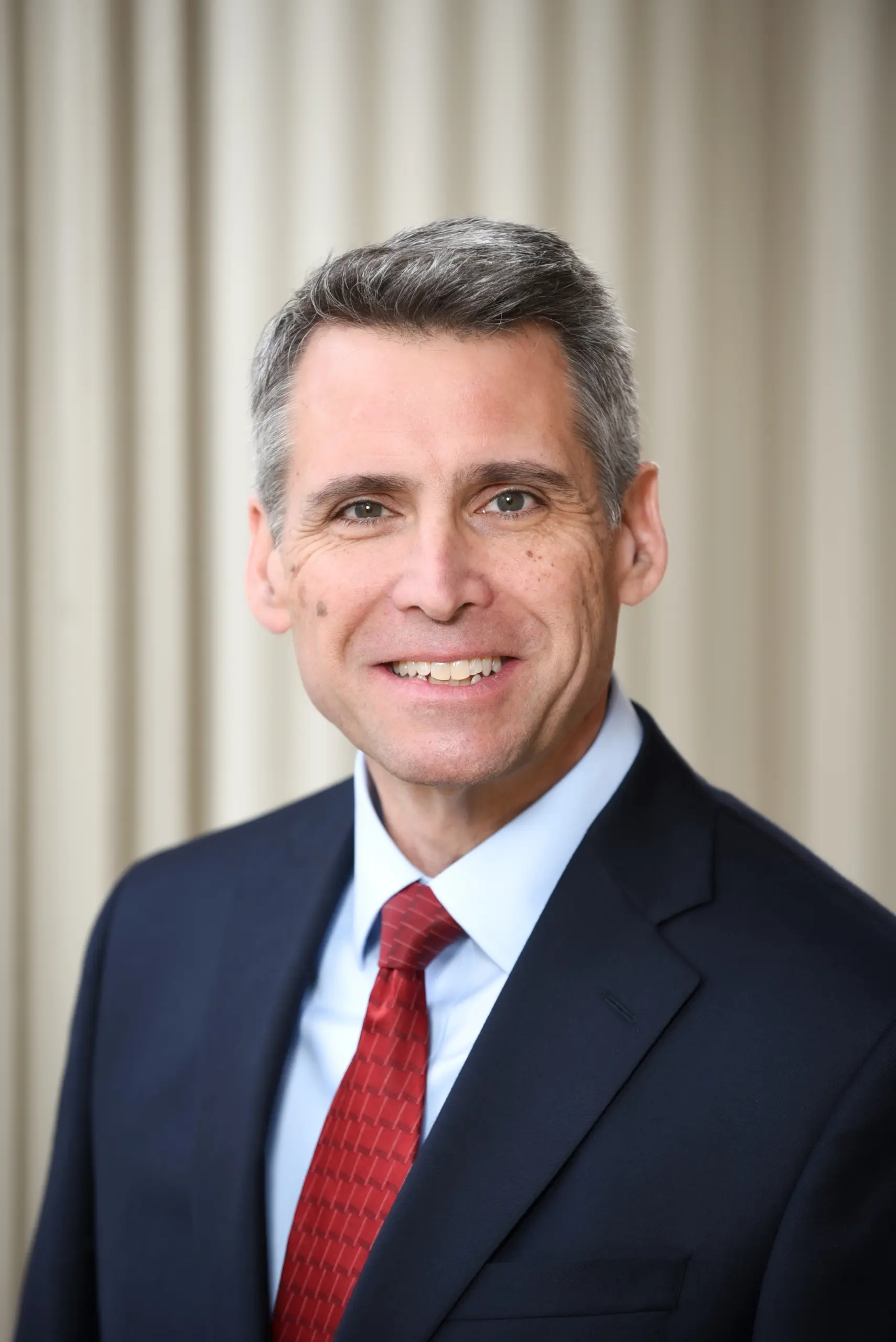 A man with short gray hair, wearing a dark suit, light blue shirt, and red tie, smiles at the camera. The background is out of focus with beige vertical lines.