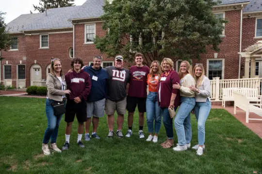 A group of nine people standing together on a grassy area in front of a brick building. They are smiling and casually dressed. Some are wearing sweatshirts and jeans. A tree and some trimmed bushes are visible in the background.