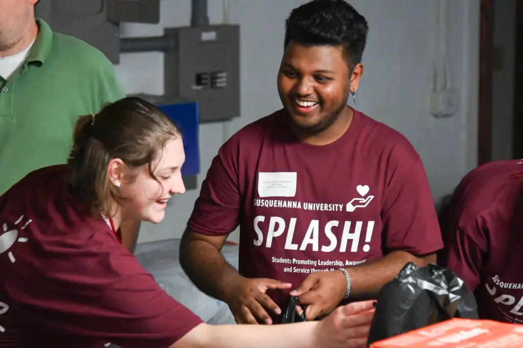 Two people wearing maroon shirts with "Susquehanna University SPLASH!" written on them are smiling and interacting while working together. They appear to be at a volunteer or community service event.