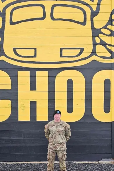 A person in military uniform stands in front of a large wall mural depicting a stylized yellow car with wings, with the word "SCHOOL" partially visible in large yellow letters.