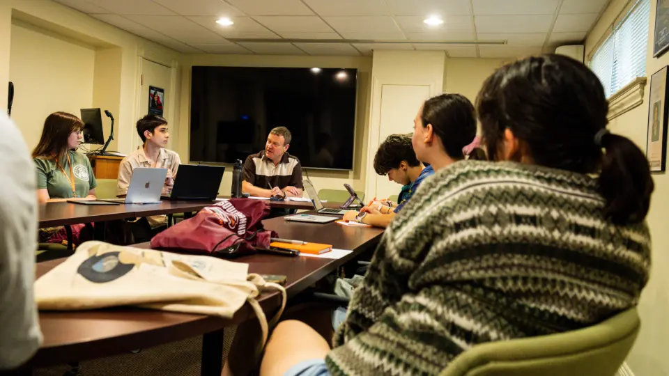 A group of people sits around a conference table in a meeting room, engrossed in a Writers Workshop. Laptops and notebooks are scattered on the table. A large television is mounted on the wall, enhancing their collaborative efforts under the room’s bright overhead lighting.