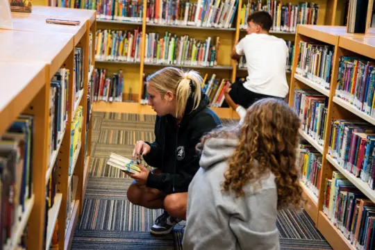 Two children search for books in a library. One, wearing a black hoodie, is crouched selecting books from a shelf. The other, in a gray hoodie, is kneeling nearby. A third child in the background, wearing a white shirt, is looking at books.