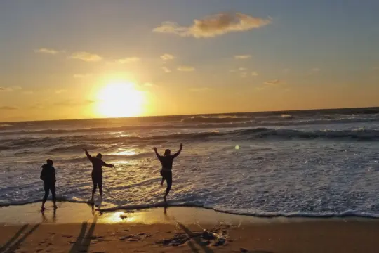 Three people on a beach enjoy a sunset over the ocean. The sun casts long shadows and highlights waves as they lap the sandy shore. The sky is partly cloudy, creating a warm, golden glow.