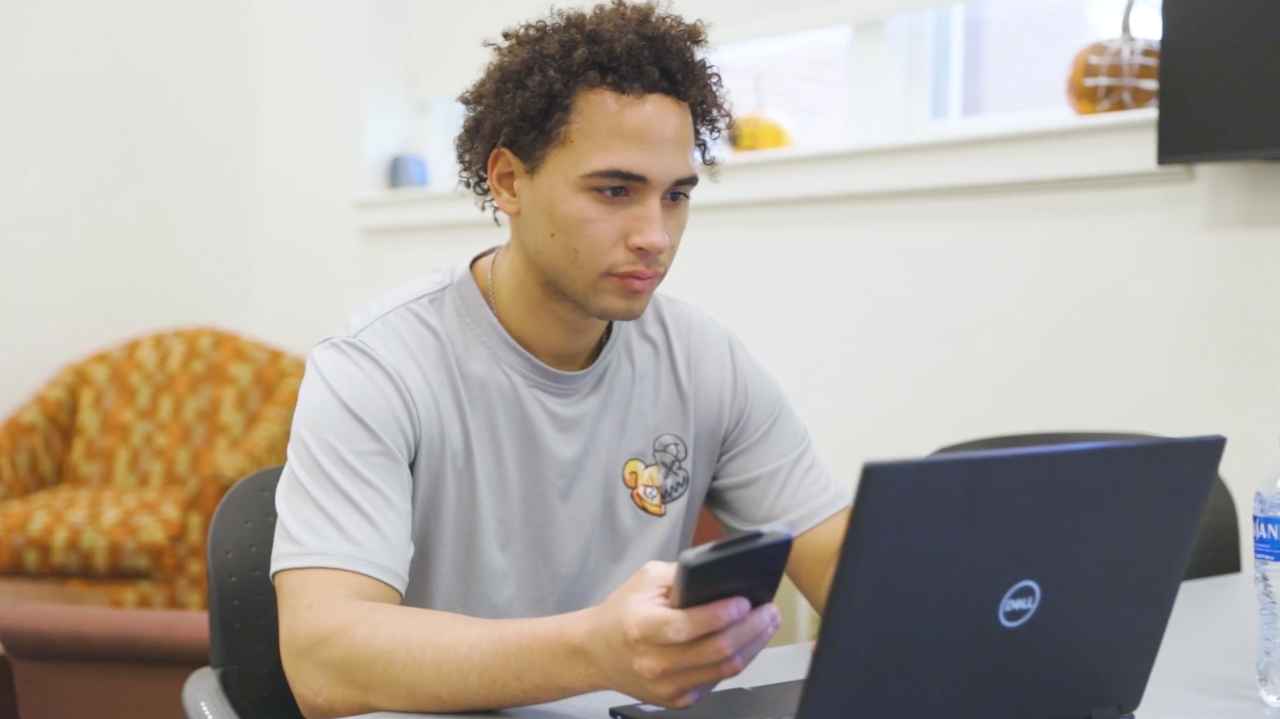 A person with curly hair is sitting at a table using a laptop and holding a smartphone. They are wearing a gray t-shirt. In the background, there's a patterned chair and windows with small pumpkins on the ledge.
