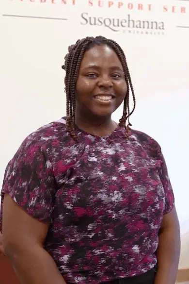 A person with braided hair smiles while wearing a purple and black patterned top. They stand in front of a wall with the words "Student Support Services" and "Susquehanna University" visible.