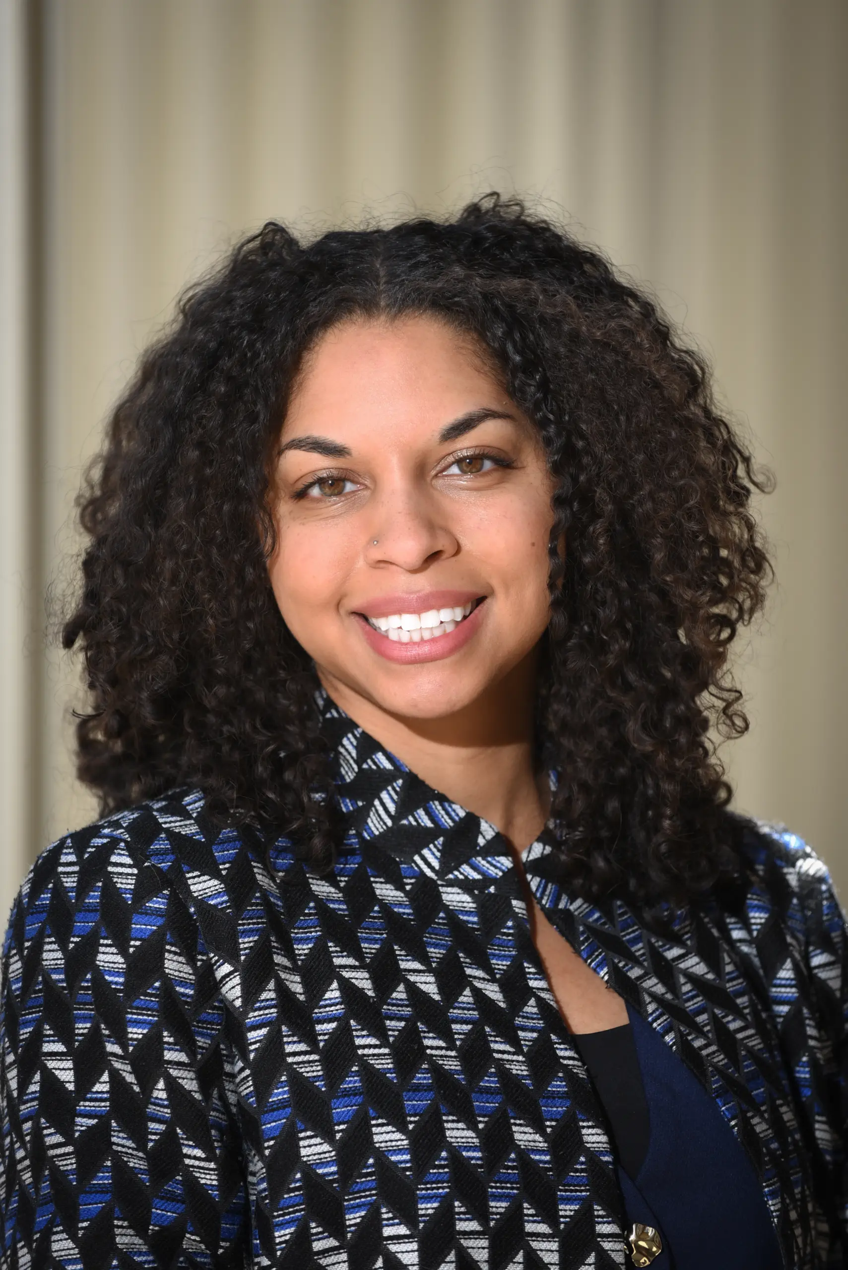 A woman with curly dark hair smiles at the camera. She is wearing a patterned blazer in black, blue, and white over a dark top, standing in front of a softly blurred beige background.