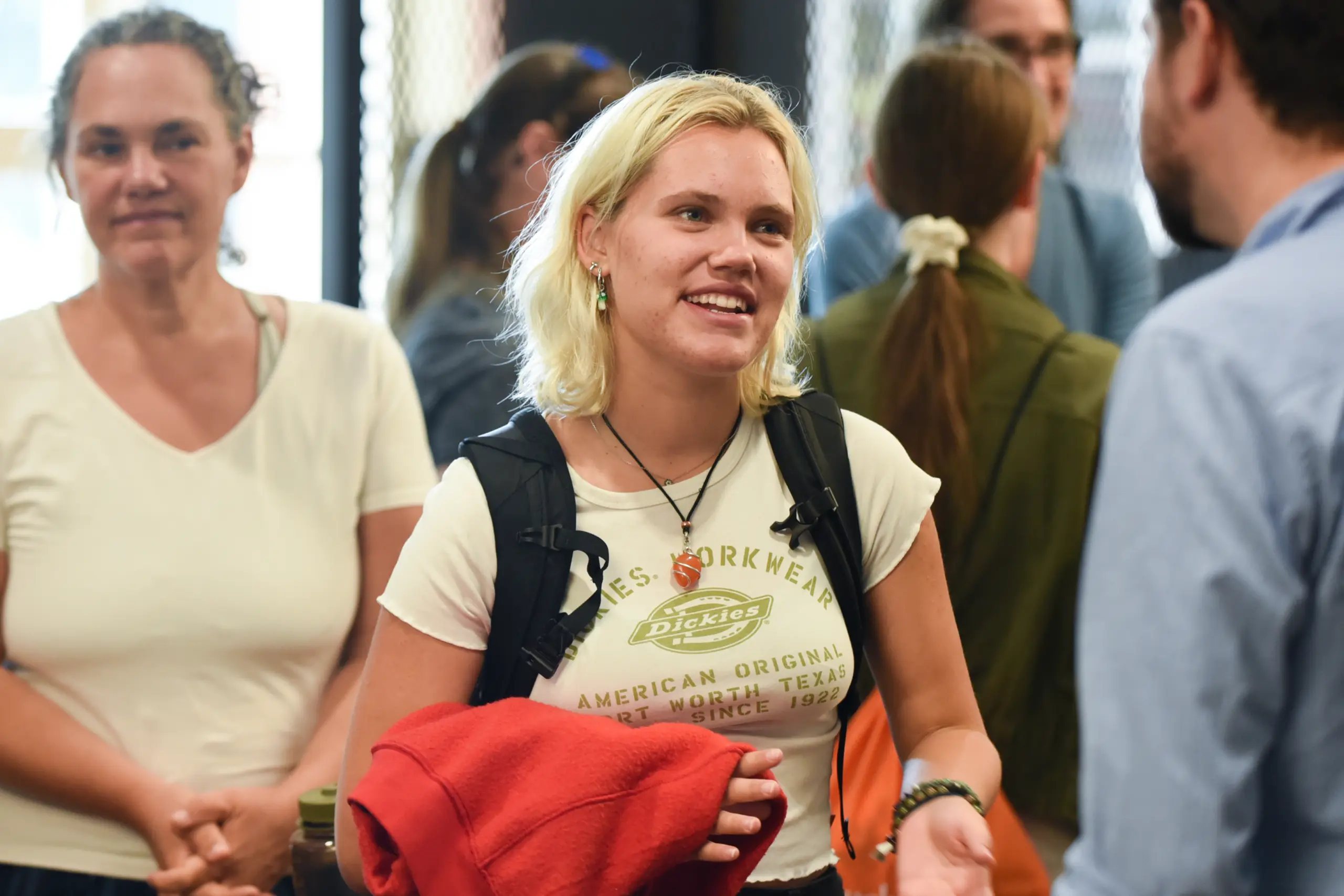 A person with blonde hair, wearing a white Dickies T-shirt and holding a red jacket, is smiling while talking to someone. Other people are in the background, casually dressed, in a social setting.