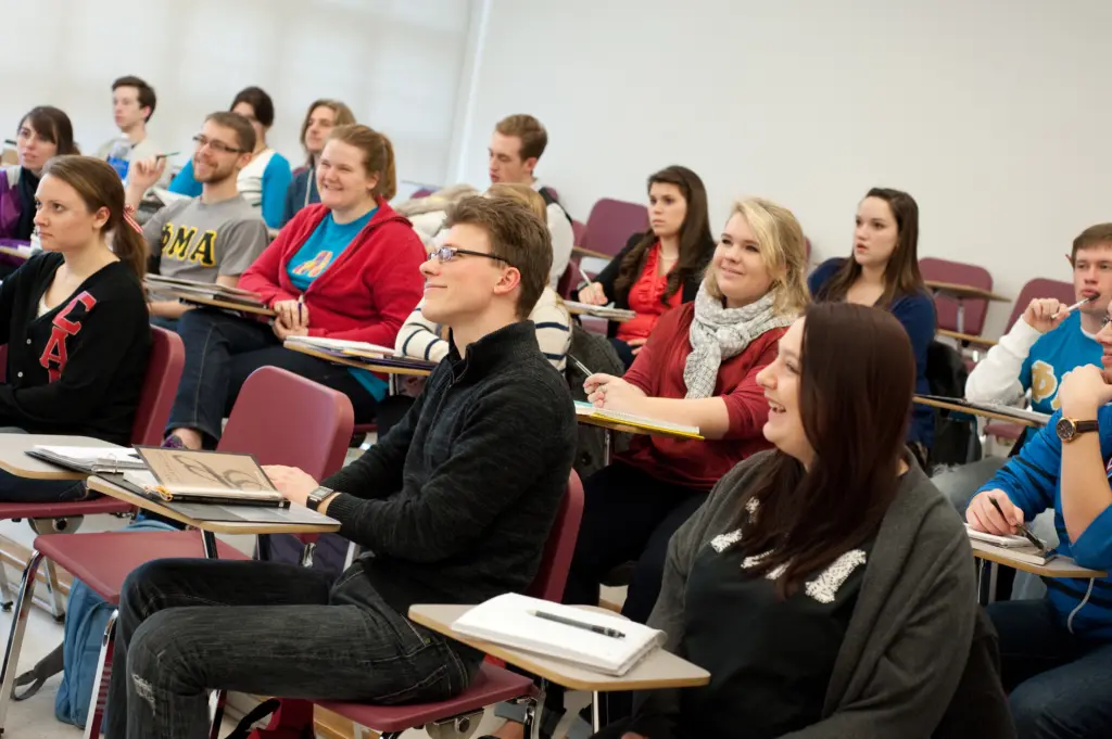A classroom filled with attentive college students, both male and female, sit at desks with notebooks open. The room has maroon chairs and wooden desks. Some students wear glasses, and the atmosphere appears focused and engaged.