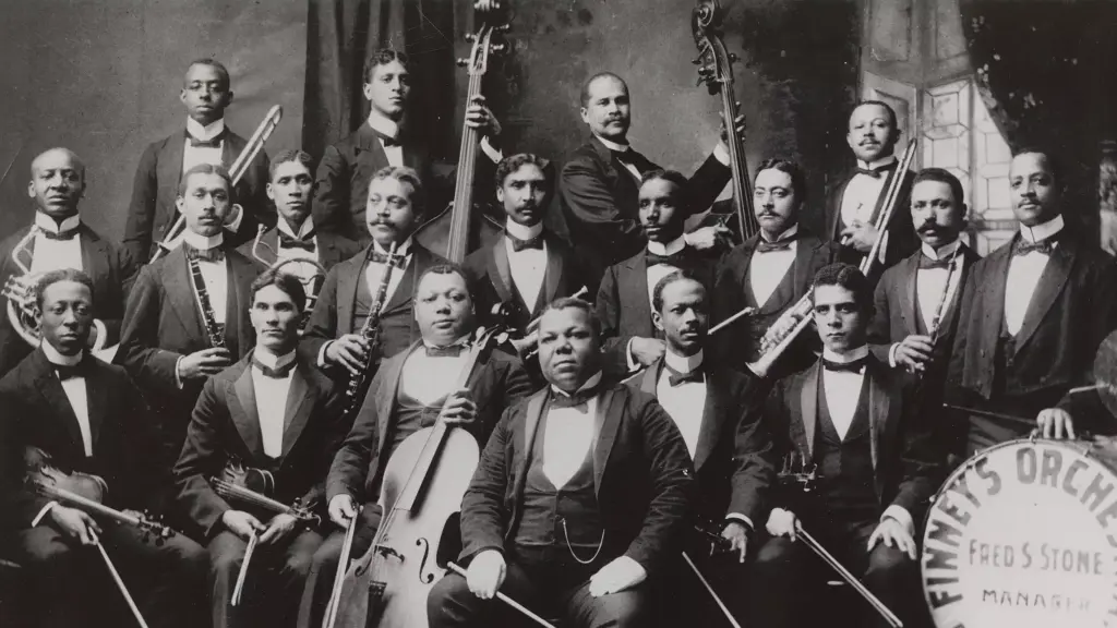 A historic black and white photo captures Finney's Orchestra, featuring sixteen men in formal attire holding their instruments. They pose in rows, with a bass drum labeled "Finney's Orchestra, Fred Stone, Manager" on the right—a nostalgic nod to the early days of music festivals.