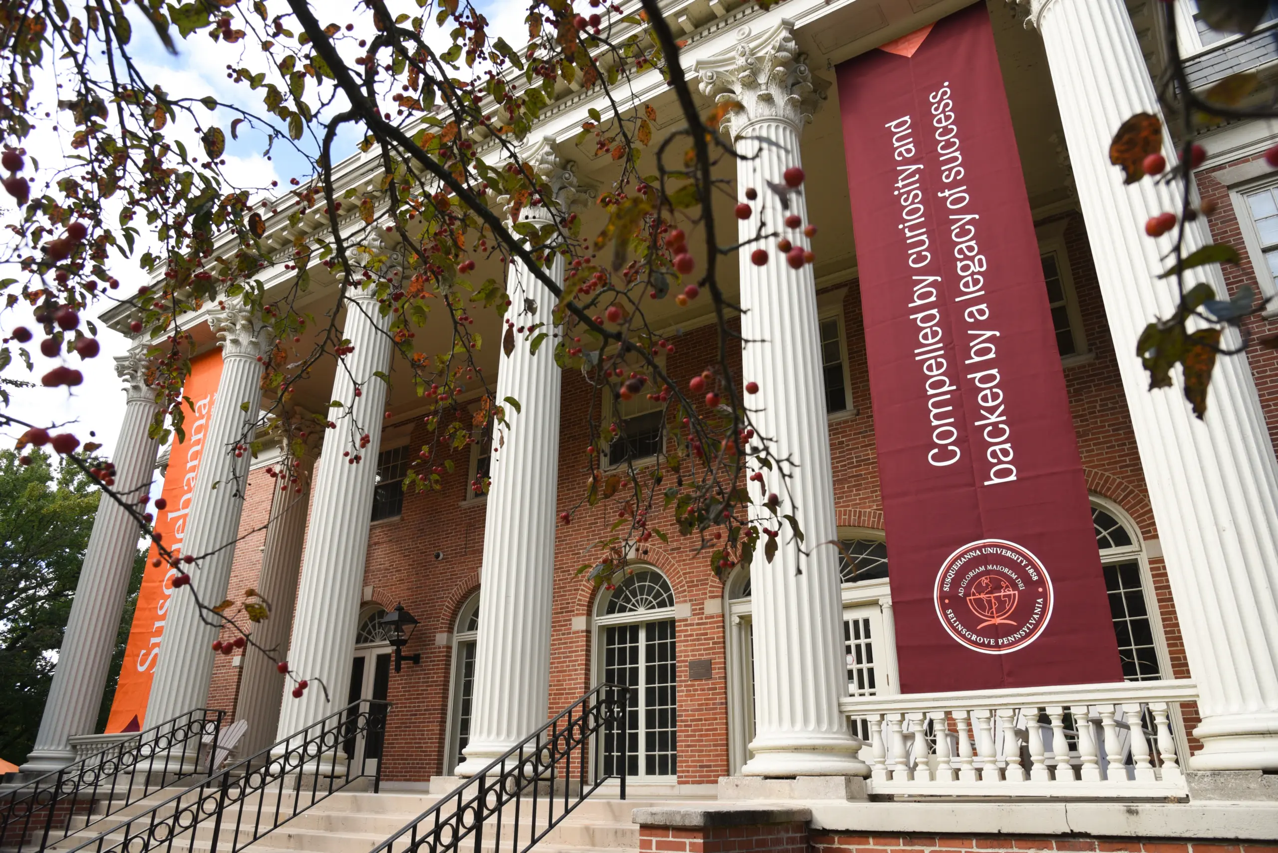 A grand building with tall white columns and red brick facade captivates viewers. One large banner reads, "Compelled by curiosity and backed by a legacy of success," reflecting what this place is all about. A smaller banner peeks out while branches with red berries hang in the foreground.