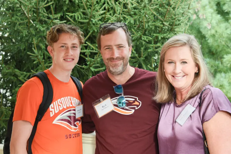 Three people attend a gathering, smiling in front of lush greenery. The person on the left wears an orange shirt, the person in the middle sports a dark shirt and glasses, and the one on the right dons a purple blouse. All proudly display their name tags.