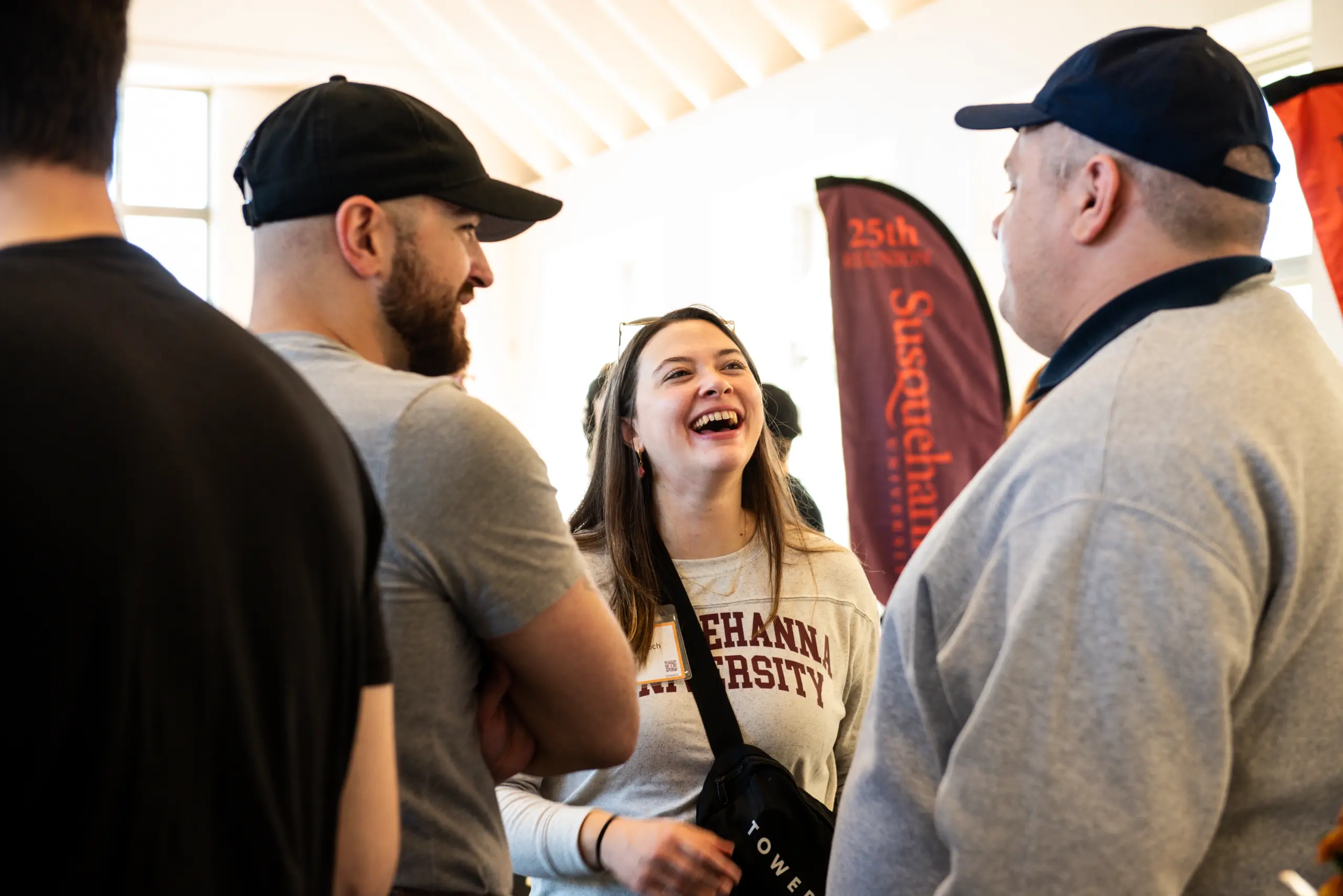 Three people gather indoors, engaged in a lively conversation. A woman wearing a "Susquehanna University" shirt attends the discussion with laughter, while two men in caps stand nearby, smiling. The ambiance is cozy with banners and soft lighting in the background.