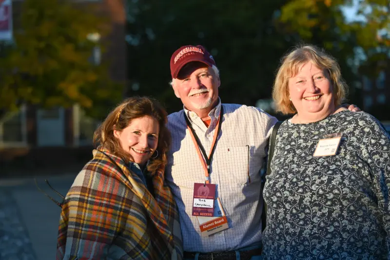 Three people are smiling outdoors. A man in the center with an all-access pass and cap has his arms around two women. The woman on the right, sporting a name badge, is an alumni. They stand in front of trees and a building bathed in warm sunlight.