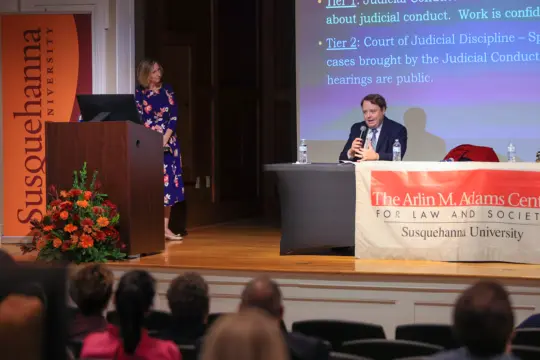 A man sits at a table speaking into a microphone on stage during the Arlin M. Adams Lecture, while a woman stands nearby. A presentation slide is projected behind them, with a Susquehanna University banner and podium visible. Audience members gather in the foreground.