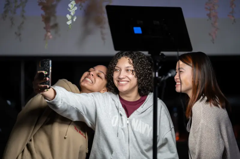Three people smiling and posing for a selfie under studio lights. They stand close together, facing the phone held by the person in the middle. Overhead, decorative flowers hang from the ceiling, adding a festive touch.
