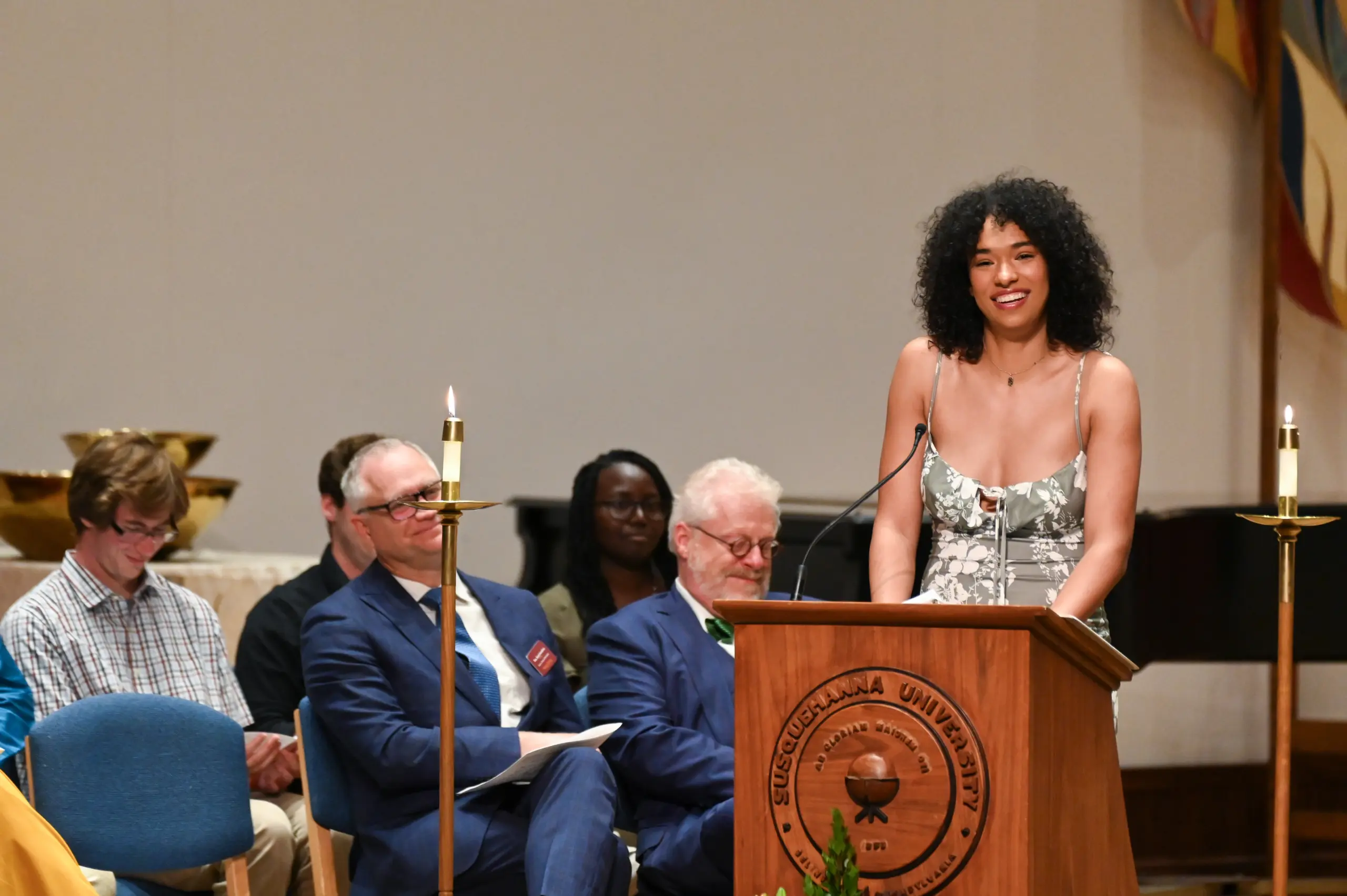 A person with curly hair, wearing a floral dress, speaks at a podium with the "Susquehanna University" emblem. Several seated people are in the background, including individuals in suits and a person with a green tie.