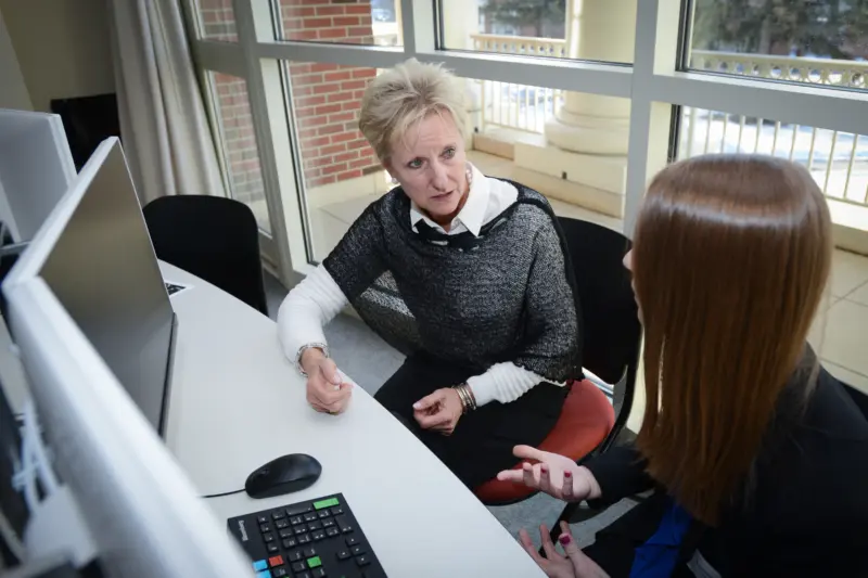Two women are seated at a desk with computers in an office. One woman is speaking while gesturing with her hands; the other listens attentively. Large windows in the background show a view outside. The environment is professional.
