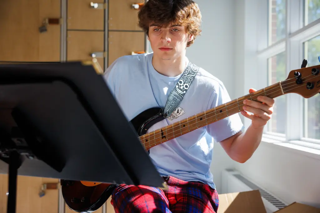 A person with curly hair is sitting and playing a bass guitar while looking at a music stand. They are wearing a light blue t-shirt and red plaid pants. Bright sunlight filters through a window, illuminating the room.