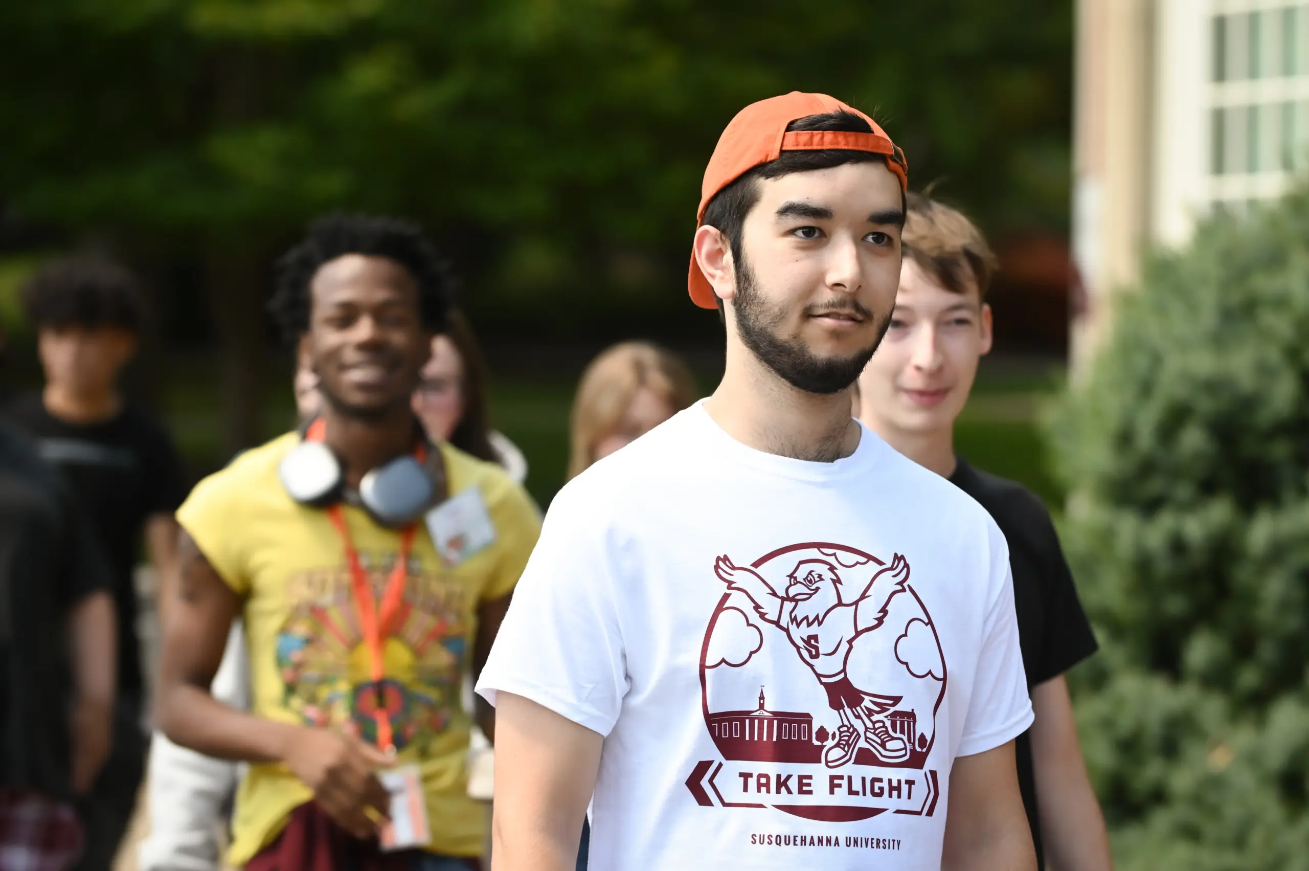 A group of young adults walking outdoors is led by someone in an orange cap and a white T-shirt with "Take Flight," reminiscent of pre-flight programs. In the slightly blurred background, one wears headphones around their neck, surrounded by lush greenery.