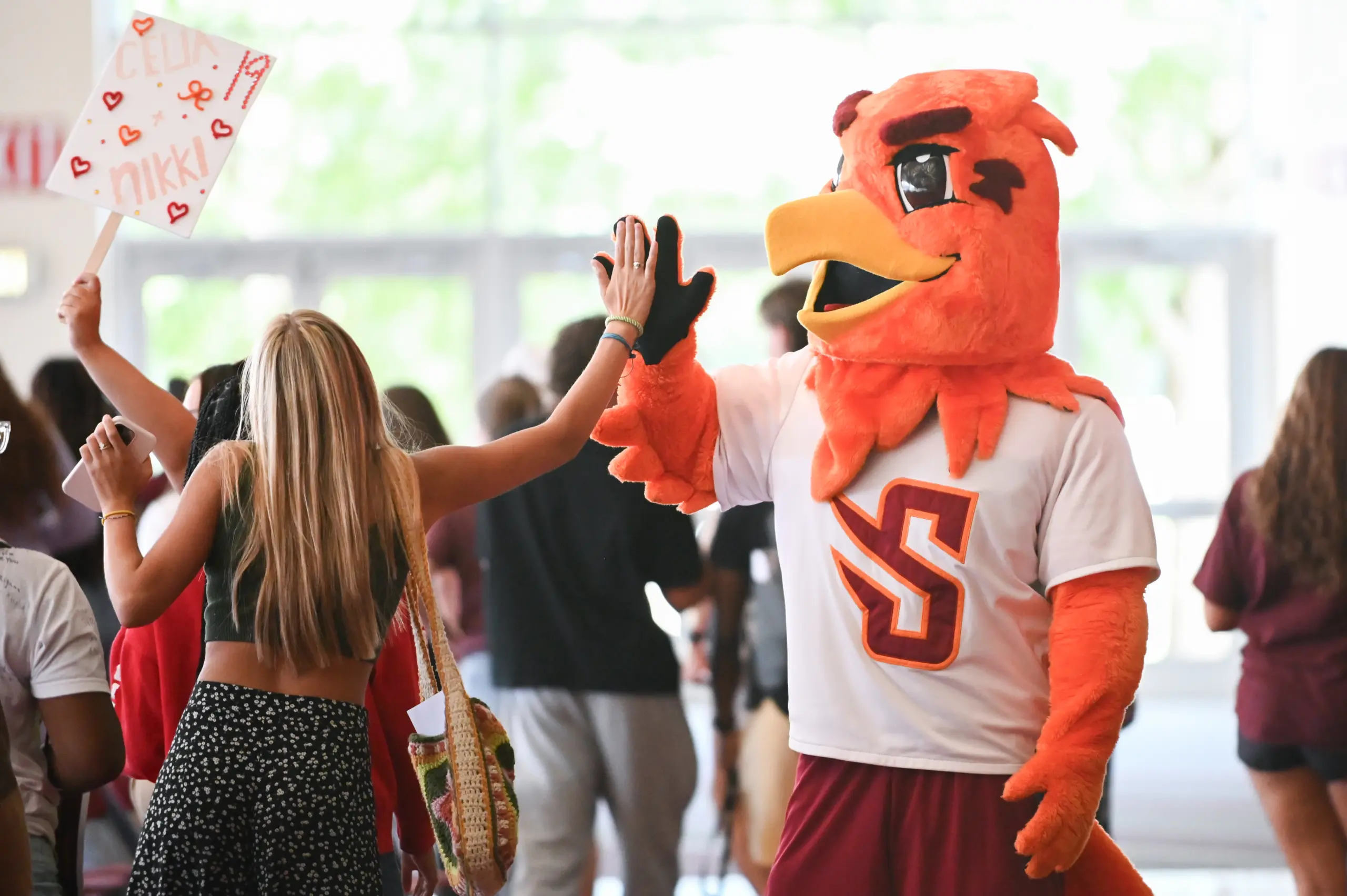 A person in a red bird mascot costume high-fives a woman with long blonde hair, celebrating her First-Year Experience. She holds a sign adorned with hearts and the name "Nikki." In the background, a crowd buzzes under bright windows.