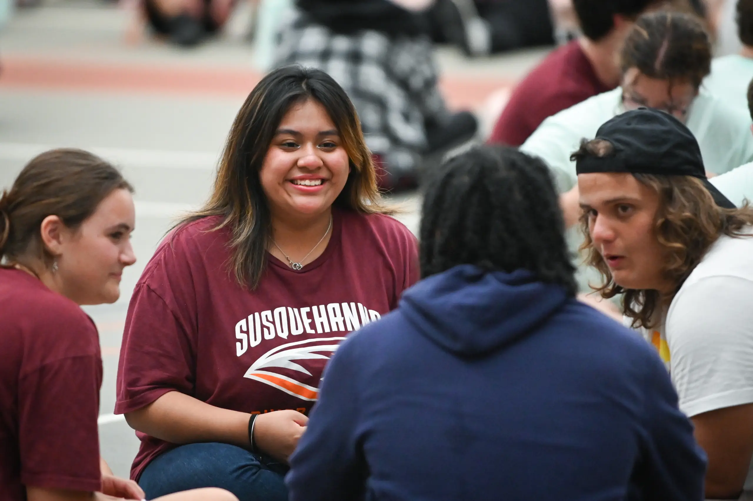 A group of young adults is gathered indoors, chatting cheerfully. At the center, one person sports a maroon "Susquehanna" T-shirt, grinning widely. The scene exudes casual ease as they enjoy the New Student Move-In excitement, with others sitting comfortably in the background.