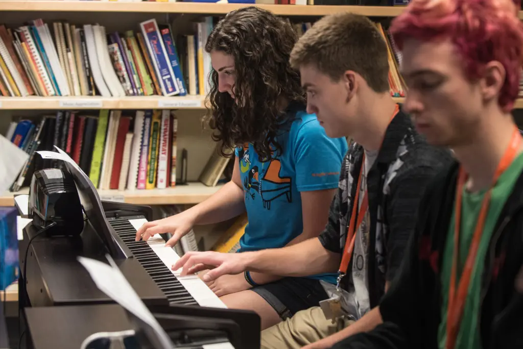Three students sitting side by side play keyboards in a classroom filled with books and musical scores. The middle student wears a blue shirt, and the others have lanyards. It's a focused music practice session.