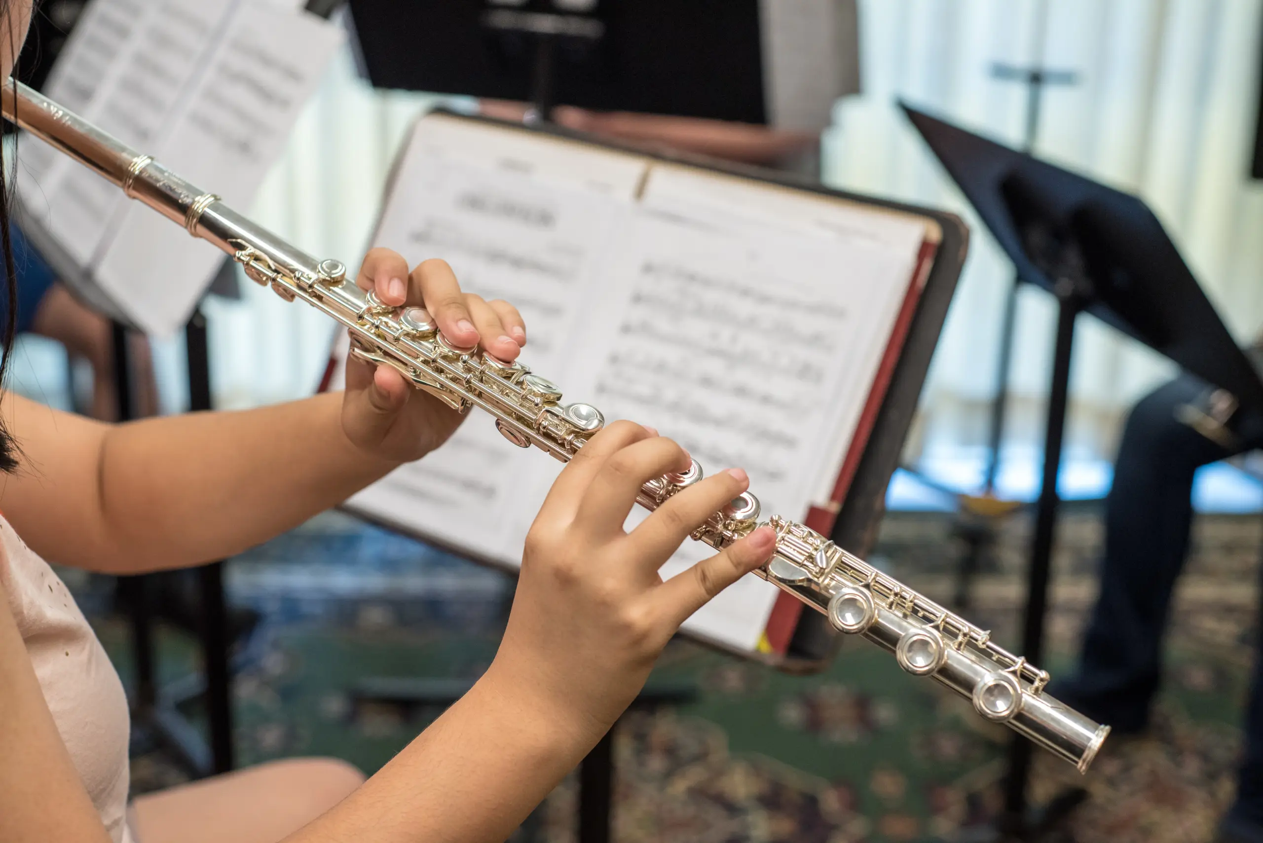 A person playing a flute, focusing on their hands and instrument. In the background, open sheet music and music stands are visible, suggesting a practice or rehearsal setting.