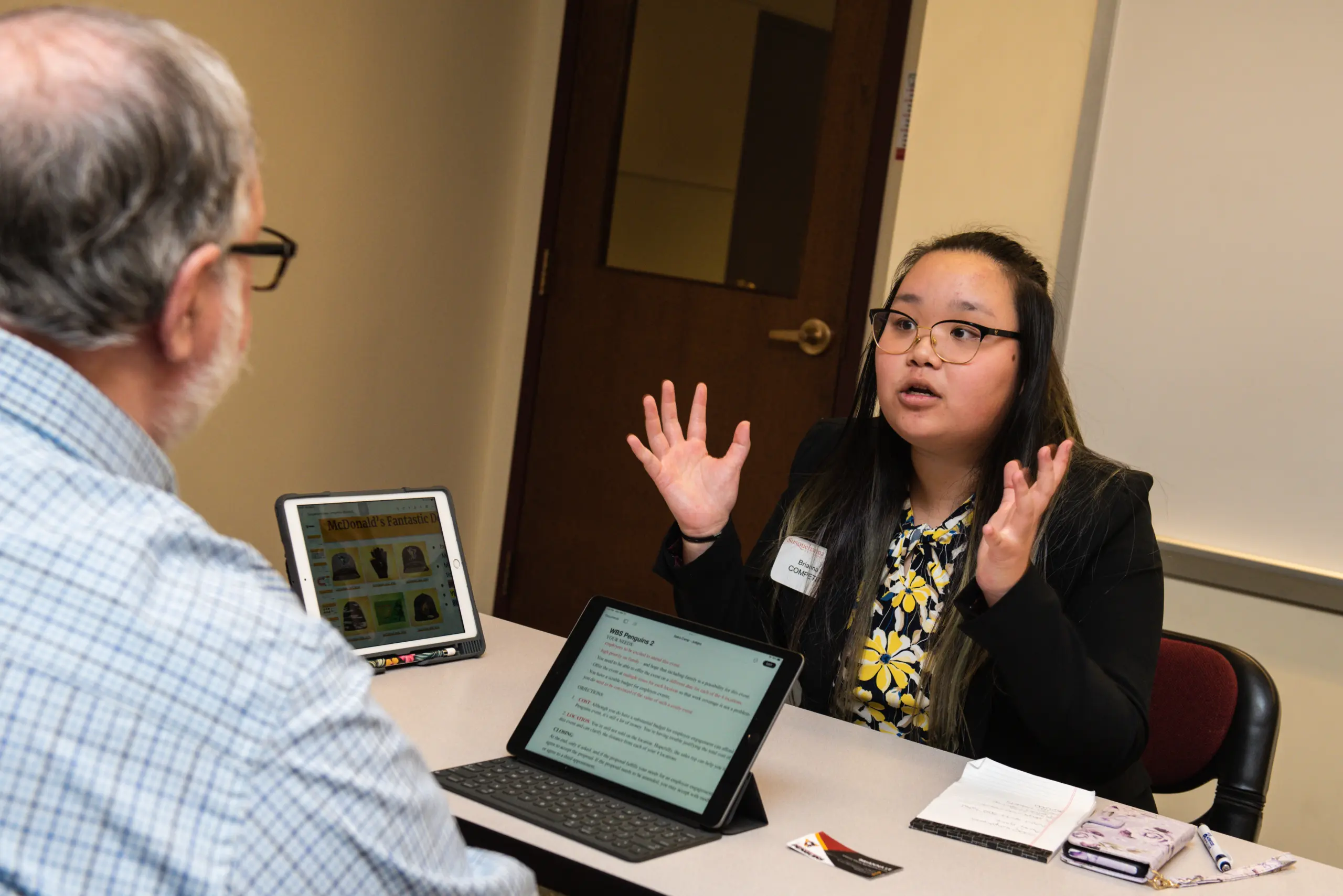 A woman with glasses is speaking animatedly with a seated man, using both hands expressively. They are at a table with tablets and papers in front of them. The room has a closed door in the background.