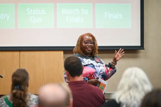 A person stands in front of an audience, speaking and gesturing with one hand. They are smiling and holding a paper. Behind them, a presentation slide with the words "Status Quo," "March to Finals," and "Finals" is displayed.