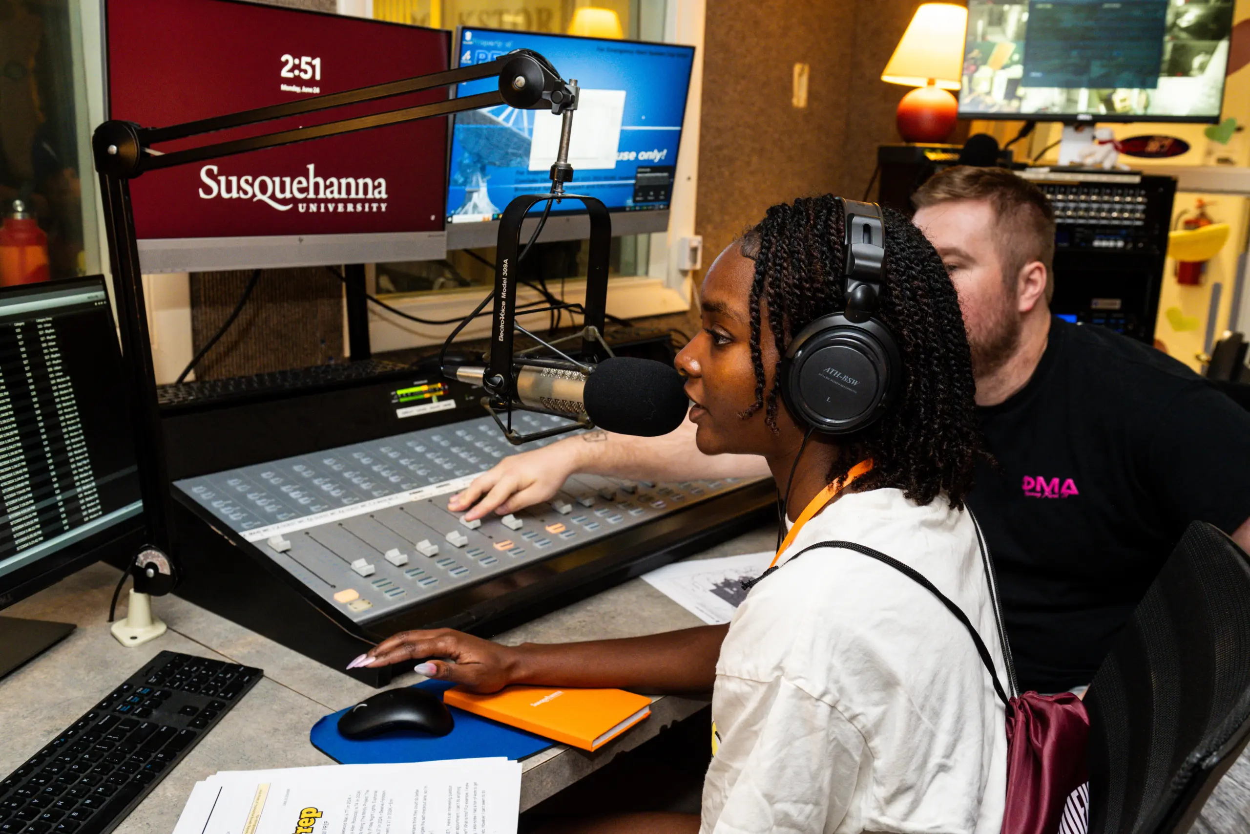 A woman speaks into a microphone in a radio studio, wearing headphones. A man sits beside her, adjusting controls on the soundboard. A computer screen displays "Susquehanna University." Papers and notebooks are on the desk.