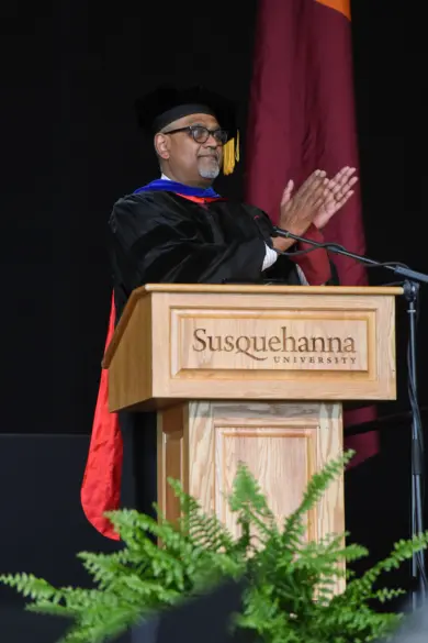 A person in academic regalia stands at a podium with "Susquehanna University" on it, clapping. A flag and a fern are visible in the background.