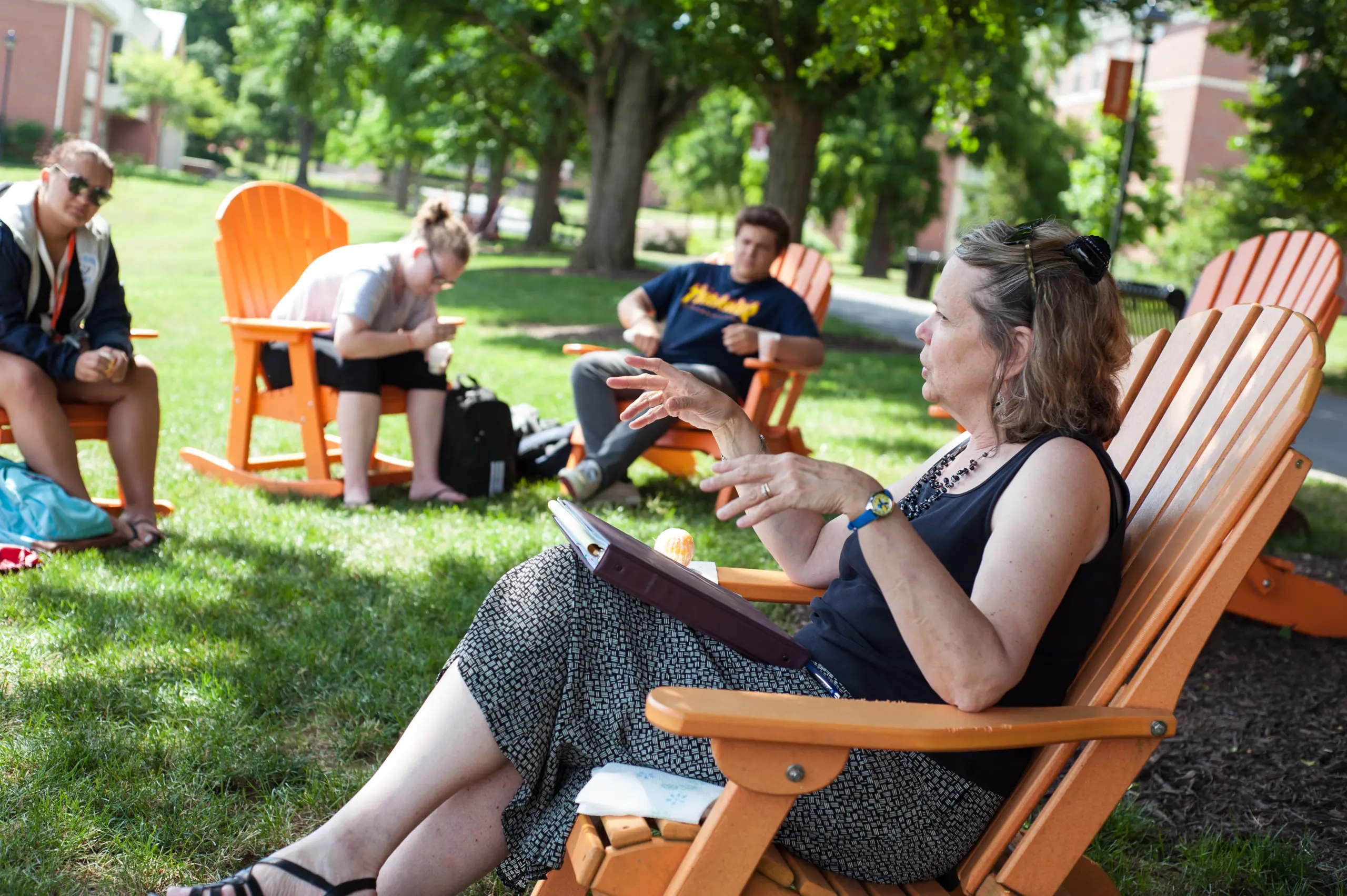 A group of people are sitting on orange Adirondack chairs outside on a grassy area. A woman gestures while holding a clipboard, and others listen. Trees and buildings are in the background, and it's a sunny day.