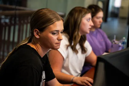 Three women focused on computer screens, seated in a row. The woman in the foreground has a black shirt, the woman in the middle has a white shirt, and the woman in the background wears a purple shirt. The setting appears to be an office.